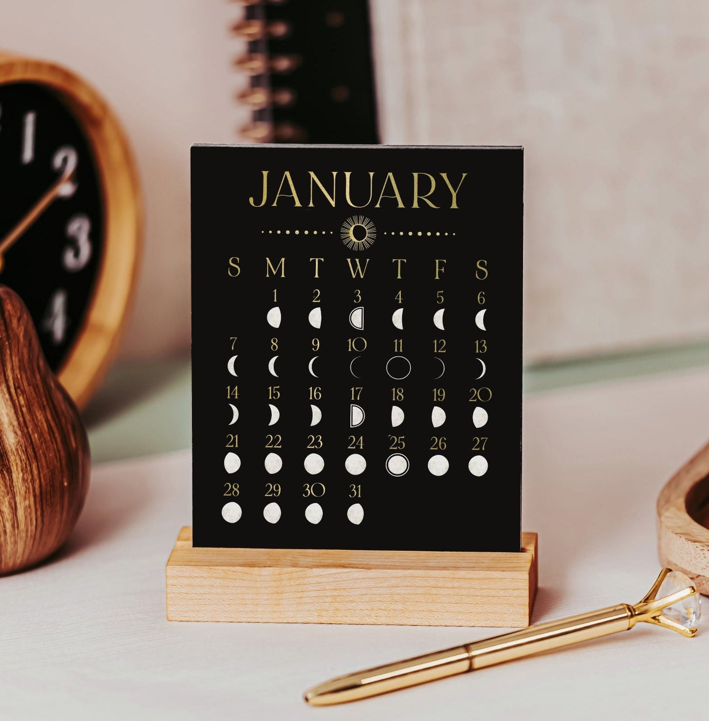 2024 full moon desk calendar with a black background and faux gold font. Each phase of the moon is featured each day of the month. It is a small desk calendar with a maple wood stand